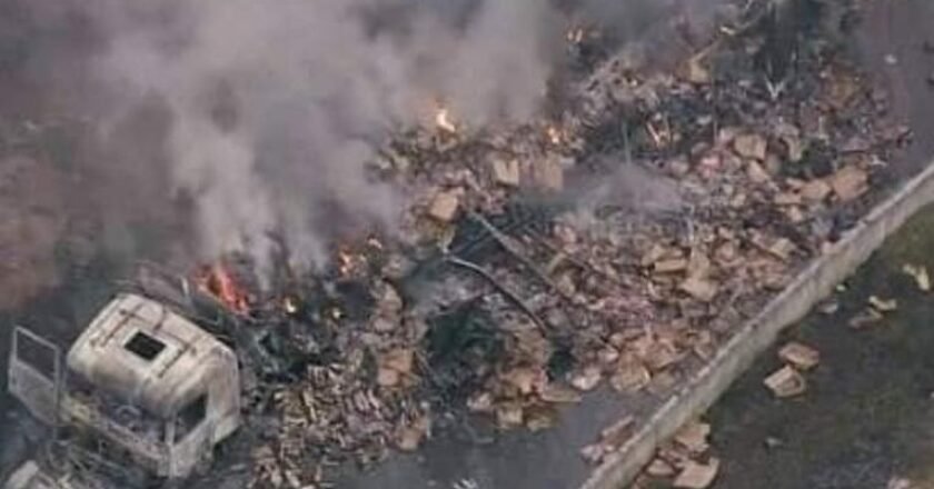 Imagem feita do alto mostra carrega incendiada, caixas com a carga espalhadas pelo asfalto. Sobre a carreta é possível ver um foco de fogo e fumaça.