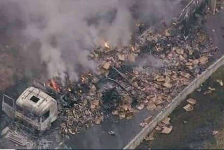 Imagem feita do alto mostra carrega incendiada, caixas com a carga espalhadas pelo asfalto. Sobre a carreta é possível ver um foco de fogo e fumaça.
