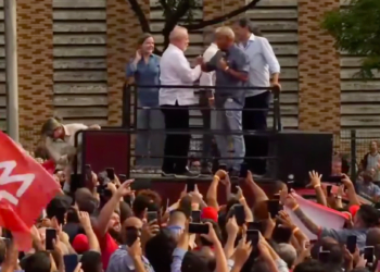 Lula em cima de palanque com outros petistas. Ele cumprimenta outro homem que está com o microfone na mão. Lula usa camisa branca. Na parte de baixo da foto aparecem mãos e celulares projetados para fazer imagens, além de uma bandeira vermelha.