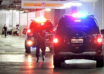 Em garagem de prédio, dos carros Polícia Federal manobram com luzes de alerta ligadas, enquanto um agente caminha ao lado das viaturas. Ao gundo, um homem e uma criança aparecem caminhando.