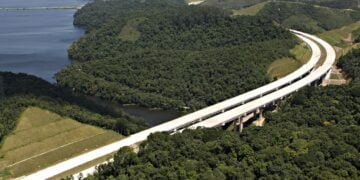 Imagem aérea mostra viaduto construído em meio a área de mata. Ao lado esquerdo está um manancial, de grande porte, que aparece sem dar dimensão exata se é um rio ou uma represa.